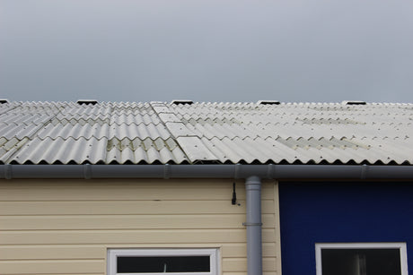 Weathered grey fibre cement roof with ridge vents above a grey PVC gutter and downpipe on a cream clad building